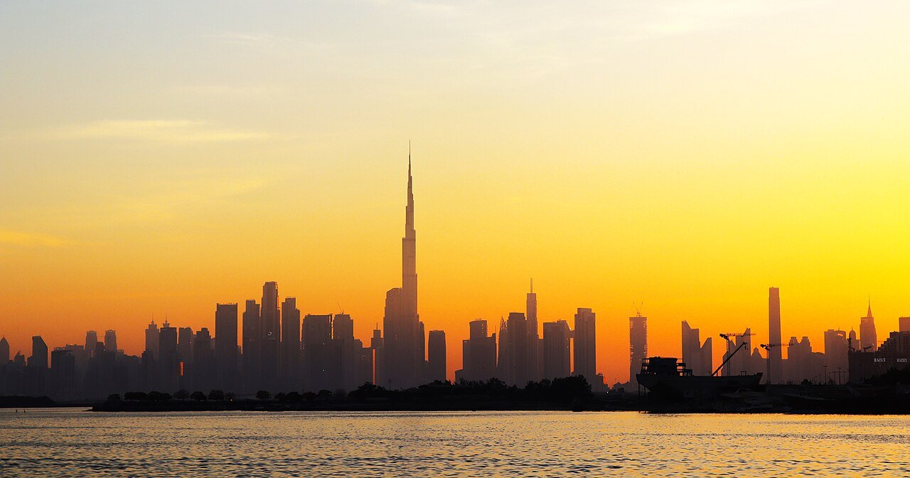 The Downtown Dubai skyline and the Burj Khalifa silhouetted against a golden sunset across the water from Dubai Creek Harbour.