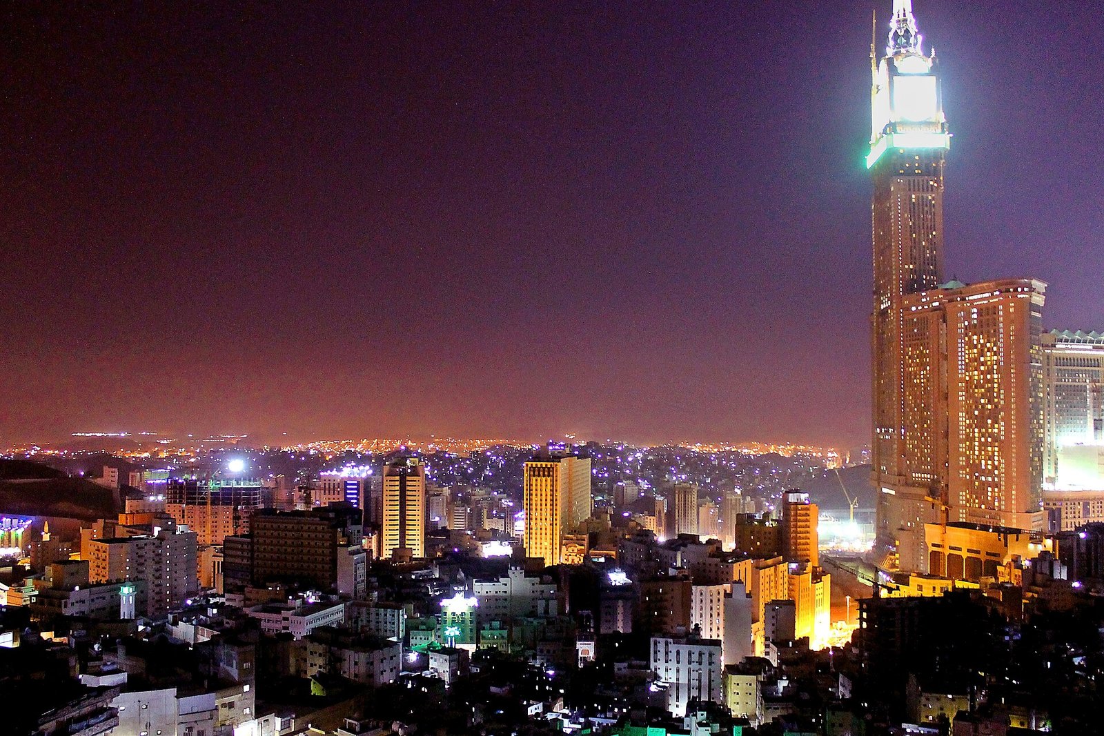 The lights of Makkah at night seen from the surrounding mountains, with the Makkah Royal Clock Tower rising at right.