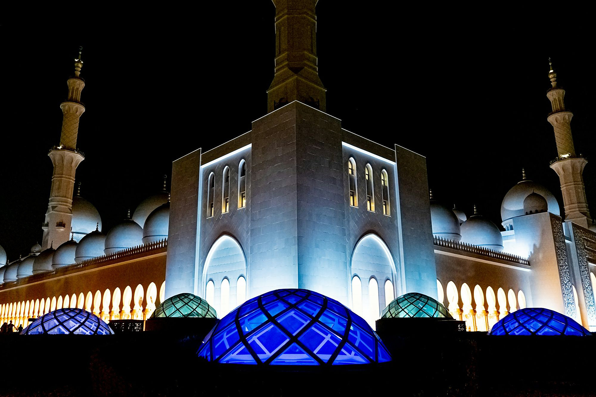 Sheikh Zayed Grand Mosque lit at night, its white domes and minarets above glowing blue glass domes
