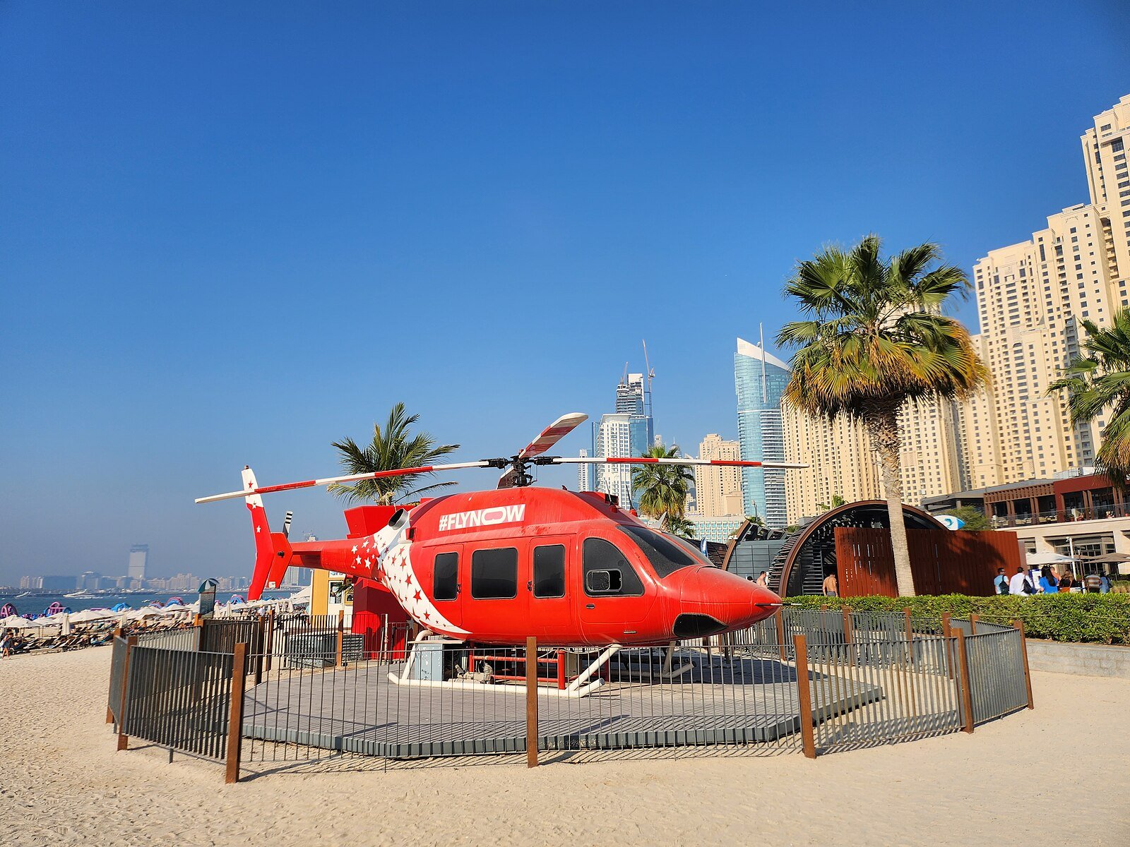 A red helicopter displayed on the sand at JBR Beach, below palms and the towers of Dubai Marina.