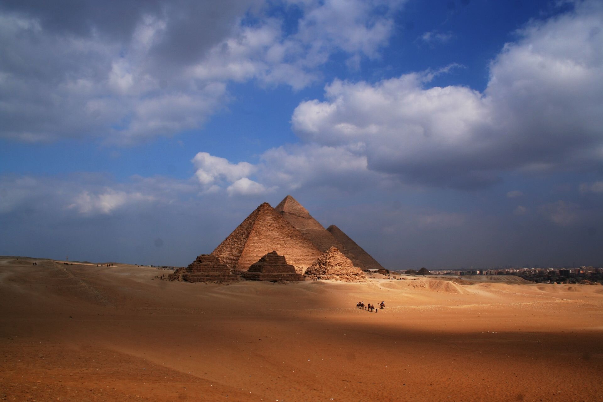 The Pyramids of Giza under a dramatic sky, with a camel caravan crossing the sand