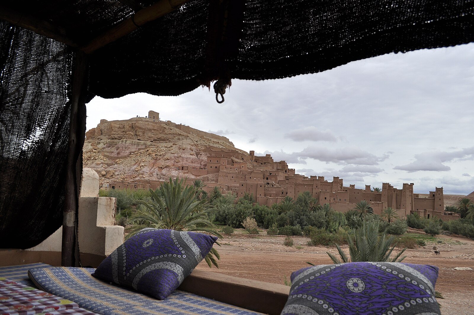 The earthen ksar of Ait Benhaddou seen from a shaded terrace with patterned cushions, near Ouarzazate.