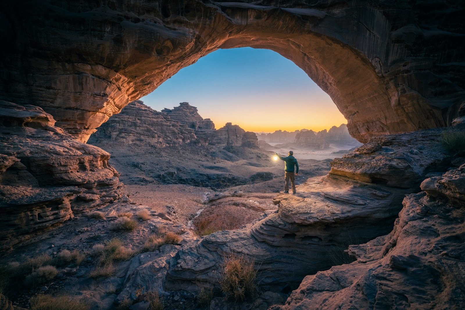 A lantern-lit traveler beneath a natural sandstone arch in the Hisma Desert at dusk