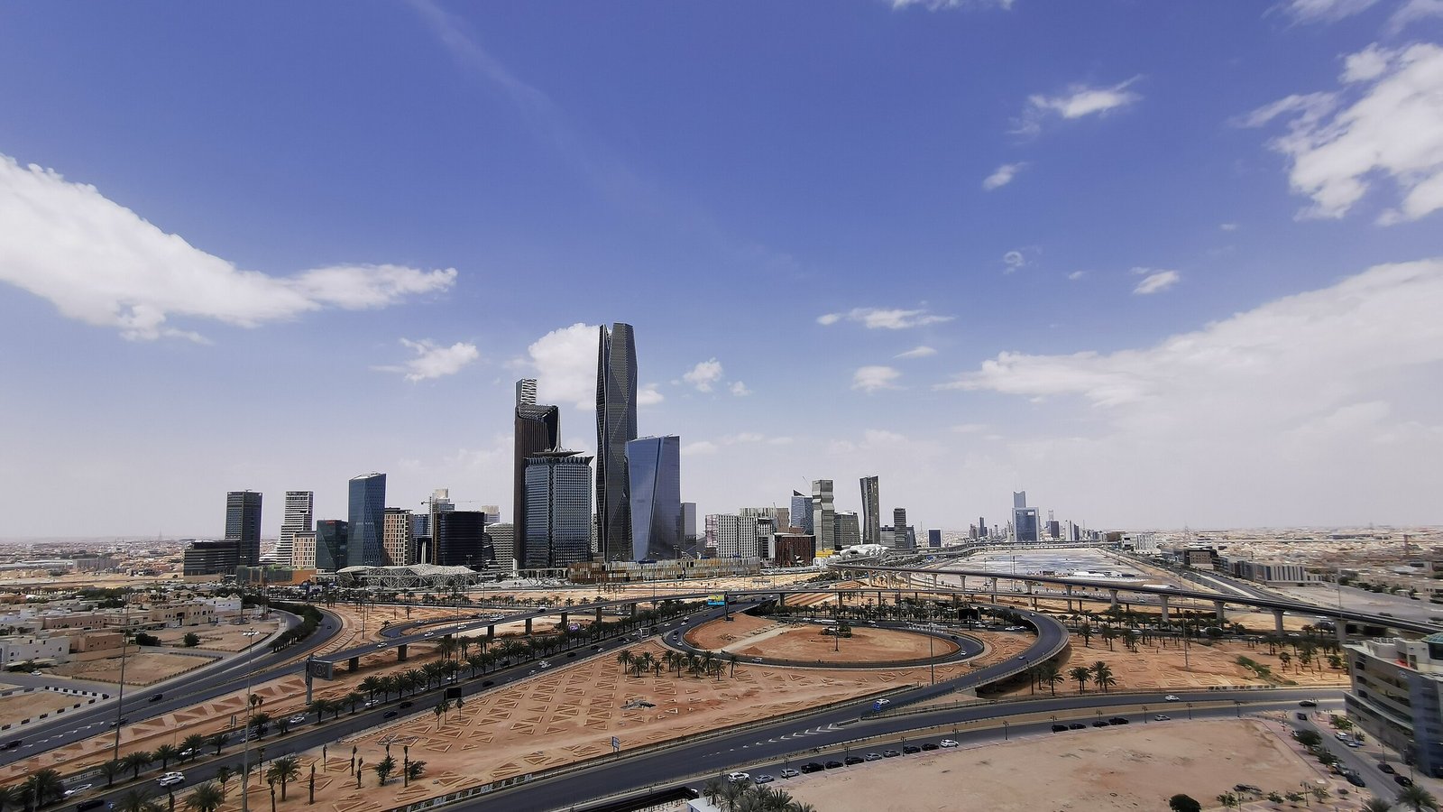 The glass towers of the King Abdullah Financial District rise above Riyadh under a bright blue sky.