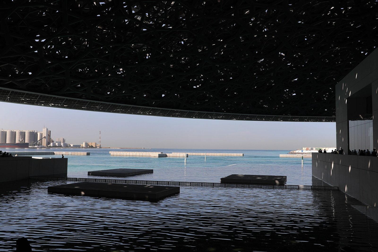Light filtering through the latticed dome of the Louvre Abu Dhabi onto the still water and the Gulf beyond.