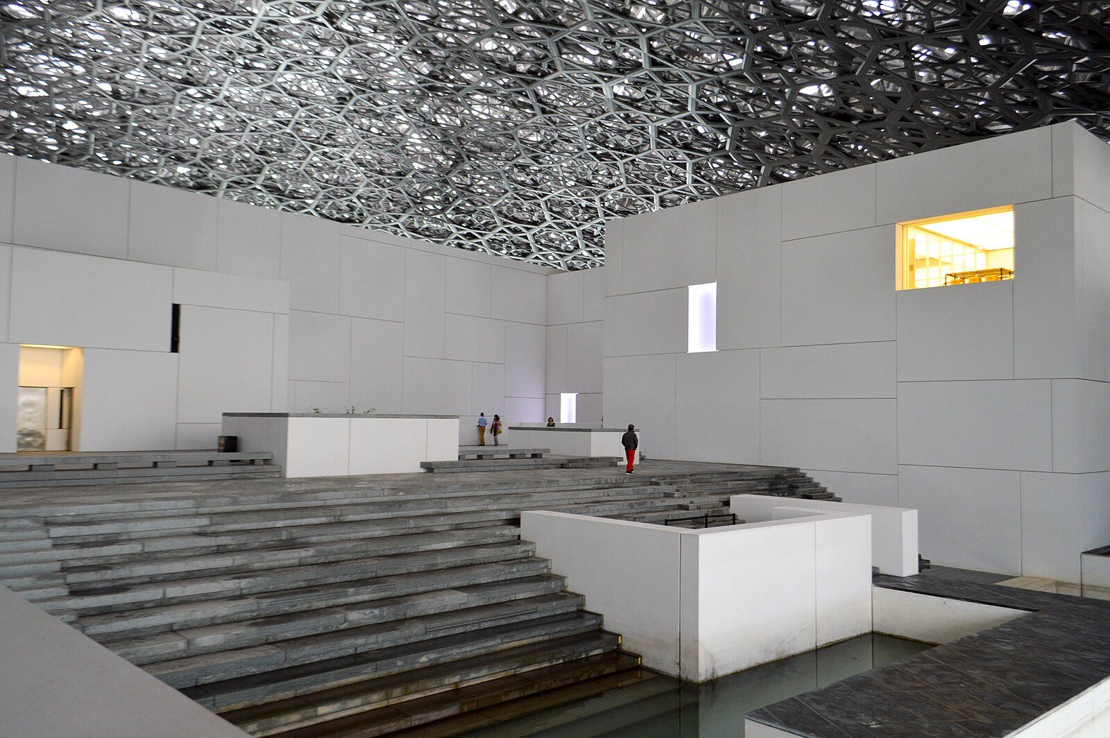 Stepped stone terraces and white gallery pavilions beneath the lace-like canopy of the Louvre Abu Dhabi