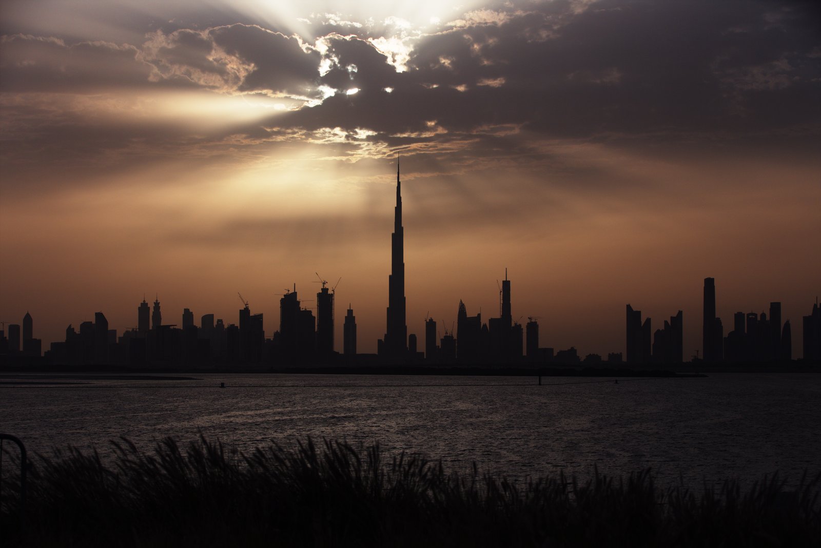 The Burj Khalifa and the Dubai skyline in silhouette beneath sunbeams breaking through the clouds.