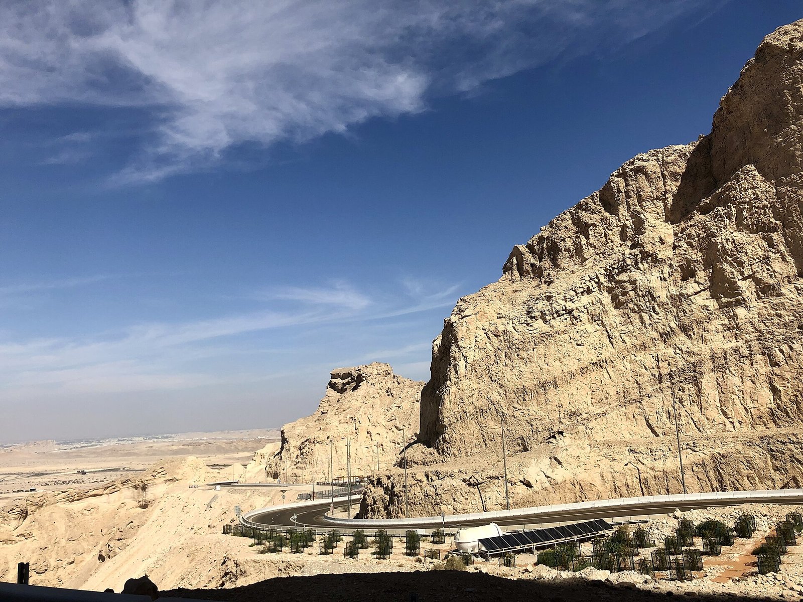 The Jebel Hafeet mountain road curves beneath sheer limestone cliffs above Al Ain under a bright blue sky.