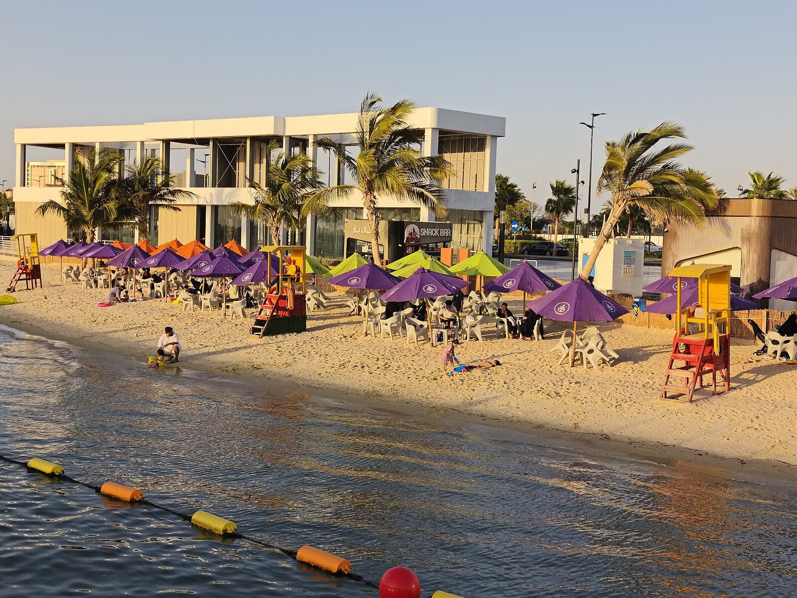 A beach club with purple parasols and palms on the South Abhur waterfront in Jeddah, in late-afternoon light.