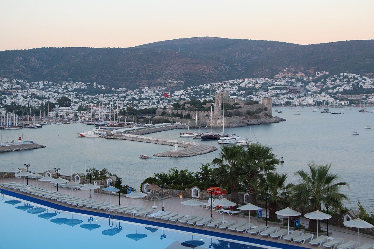 A resort pool terrace above Bodrum harbor at dusk, with the Castle of St. Peter and the town beyond.