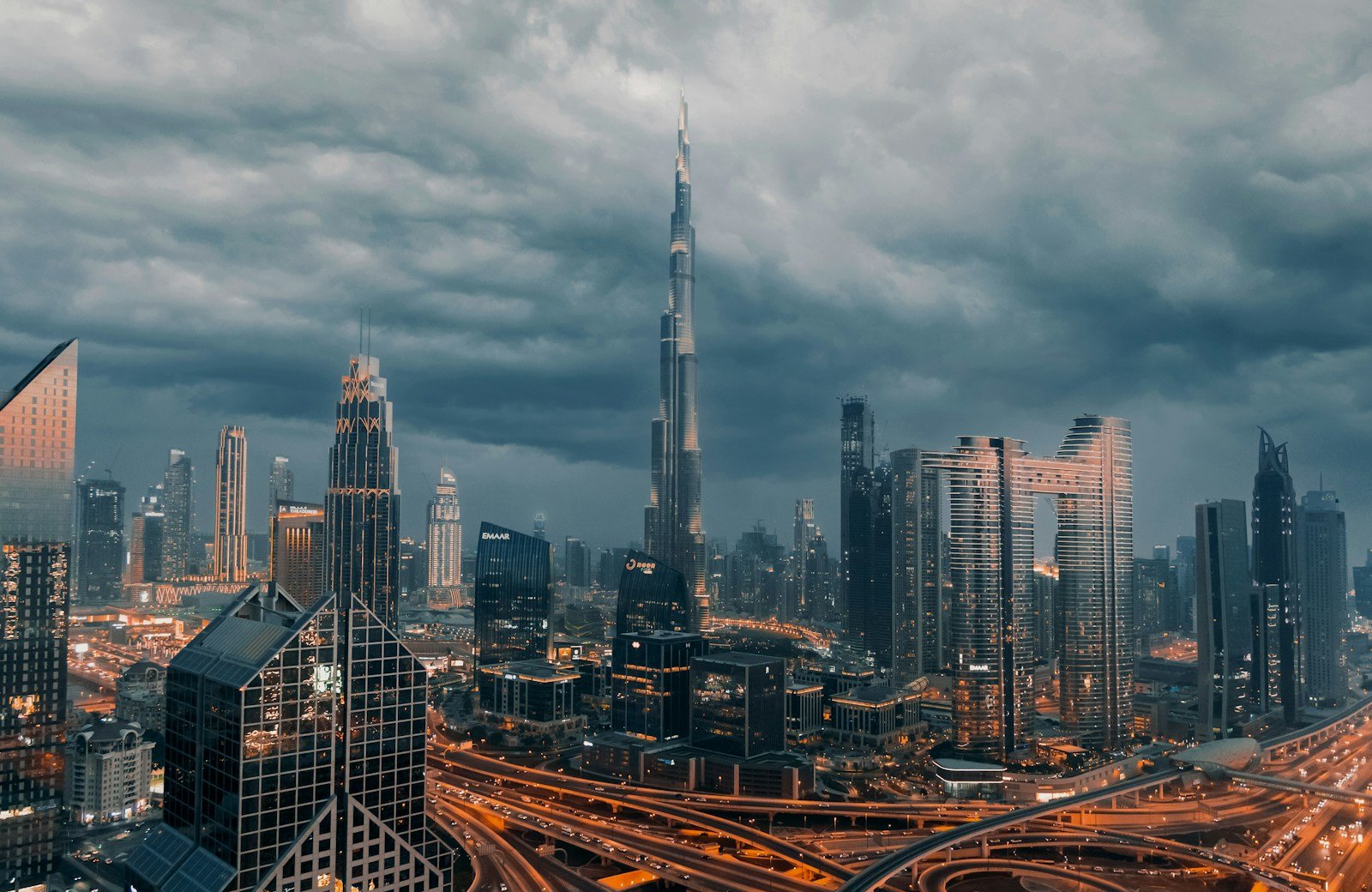 The Burj Khalifa and the lit towers of Downtown Dubai beneath a dramatic sky of gathering clouds at dusk.