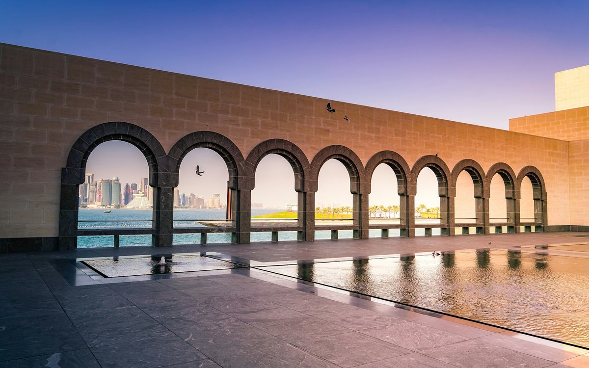 The arched loggia of the Museum of Islamic Art frames the Doha skyline across the bay at dusk.