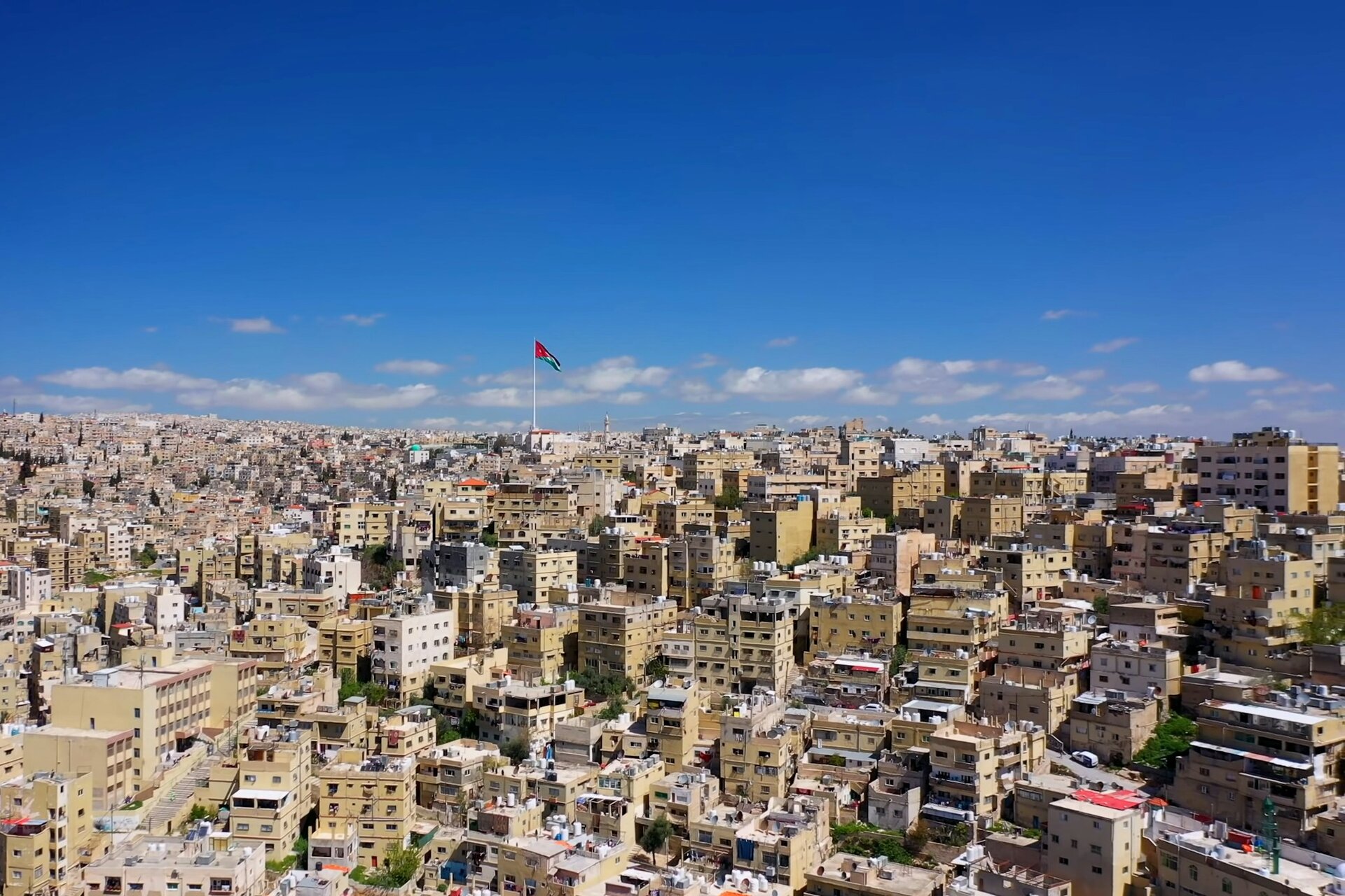 Amman's white stone hills under a deep blue sky, the Jordanian flag flying above
