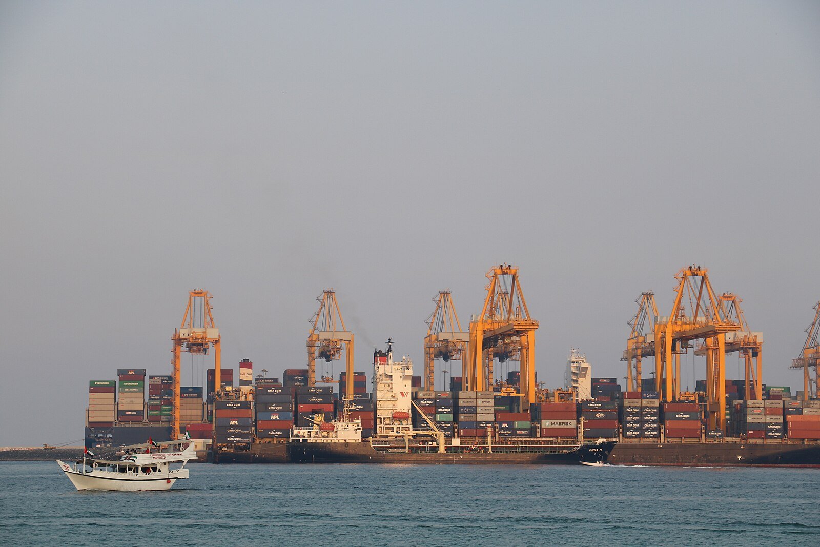 A container ship beneath the gantry cranes of the Port of Fujairah, with a traditional dhow passing in the foreground.
