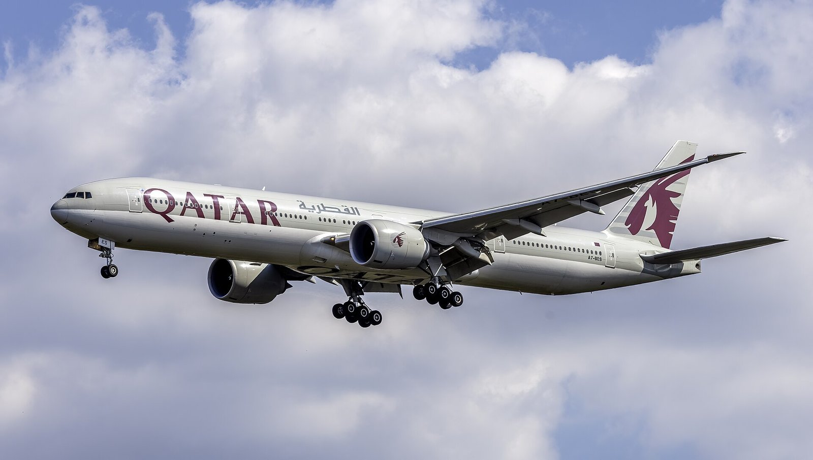 A Qatar Airways Boeing 777 on final approach to Washington Dulles under a bright cloud-filled sky.