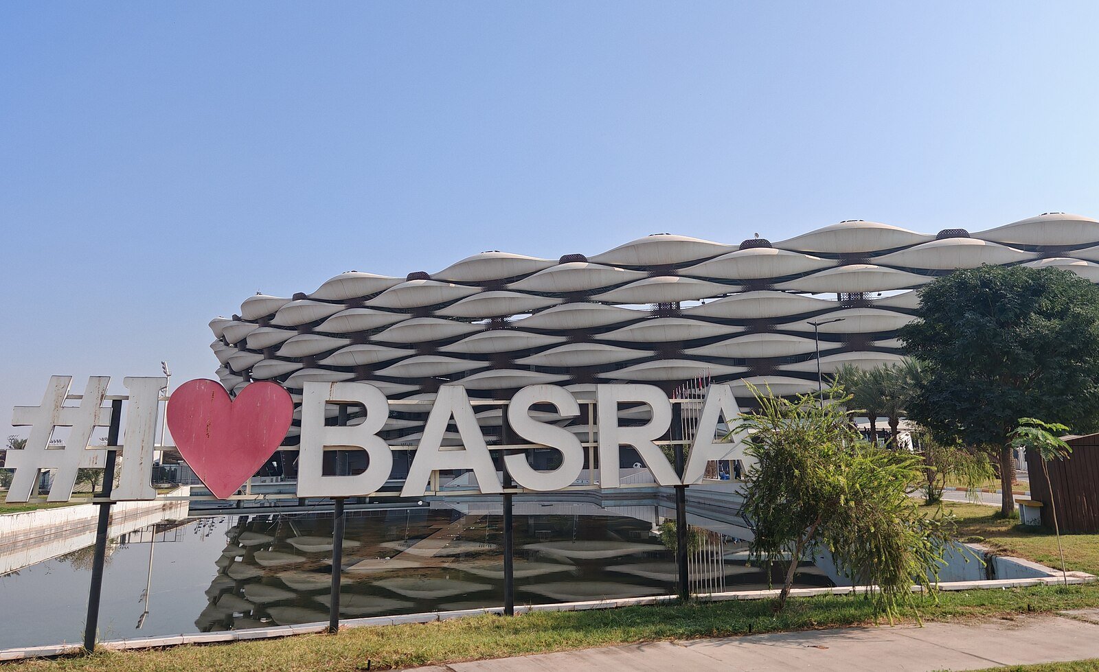 The palm-inspired lattice of Basra International Stadium reflected in a pool behind the I Love Basra sign.