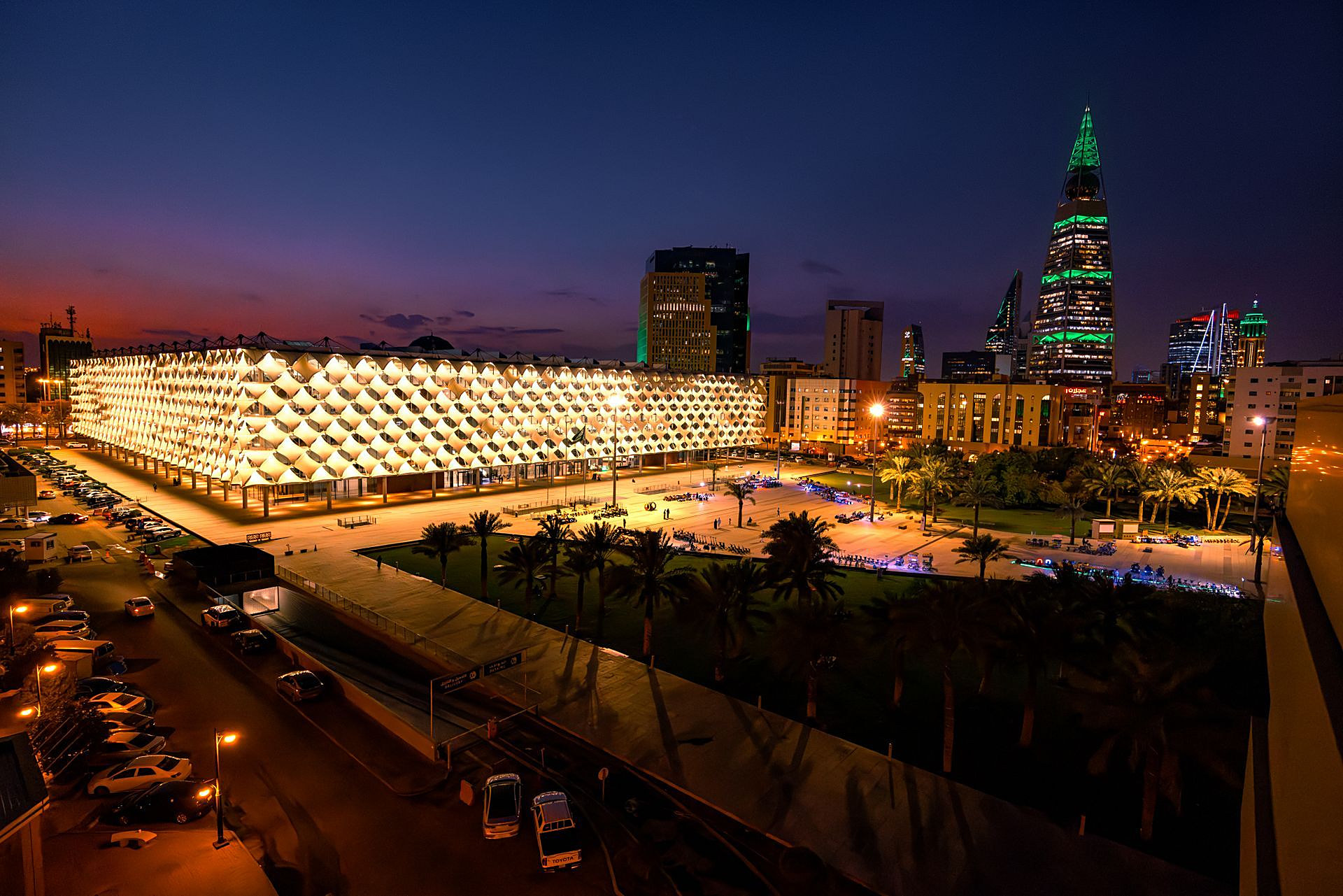 The glowing lattice facade of King Fahd National Library at night, with Al Faisaliah Tower lit green beyond