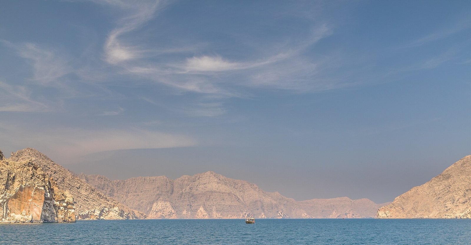 A lone dhow crosses the calm, turquoise waters of a fjord between the mountains of the Musandam Peninsula, Oman.