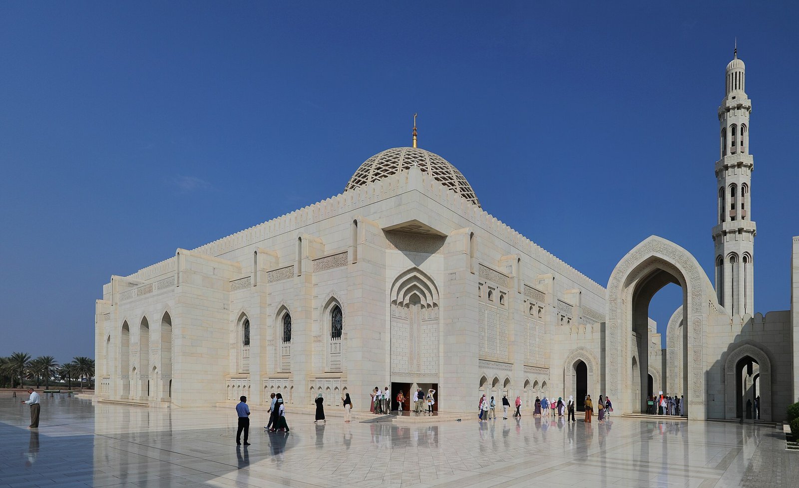 The white marble prayer hall and minaret of the Sultan Qaboos Grand Mosque in Muscat under a deep blue sky.