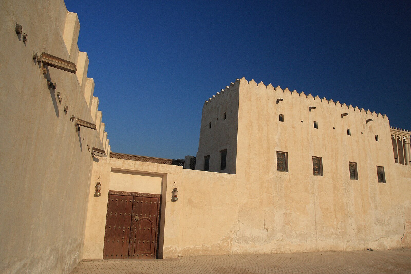 Sand-colored coral-stone walls and a carved wooden door in the Sharjah Heritage Area beneath a deep blue sky.