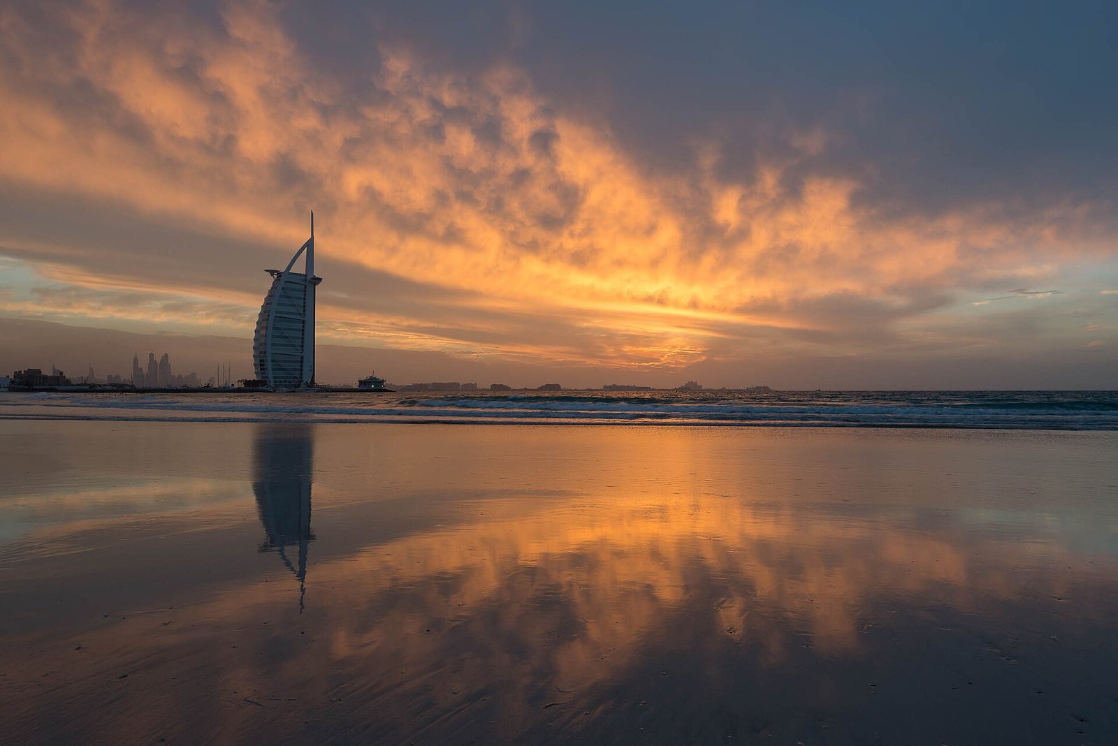 The Burj Al Arab silhouetted against a fiery sunset sky, mirrored on the wet sand of Jumeirah Beach.