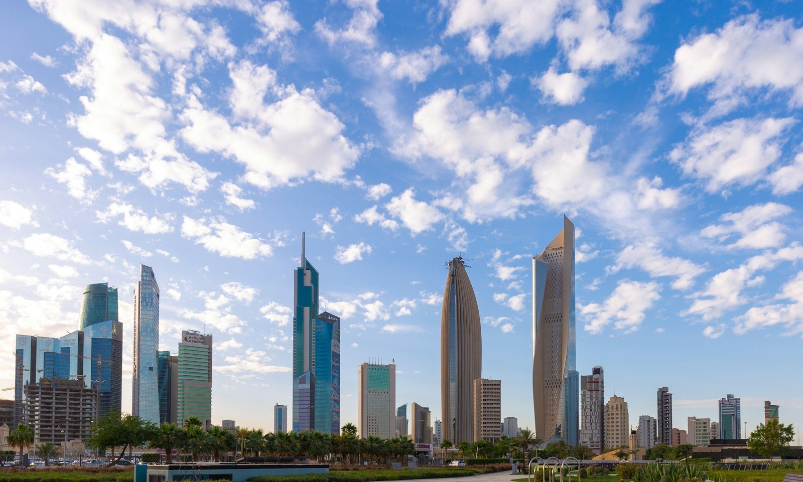 The towers of Kuwait City, led by the sculpted Al Hamra Tower, rising beyond Al Shaheed Park under a sky of scattered clouds.