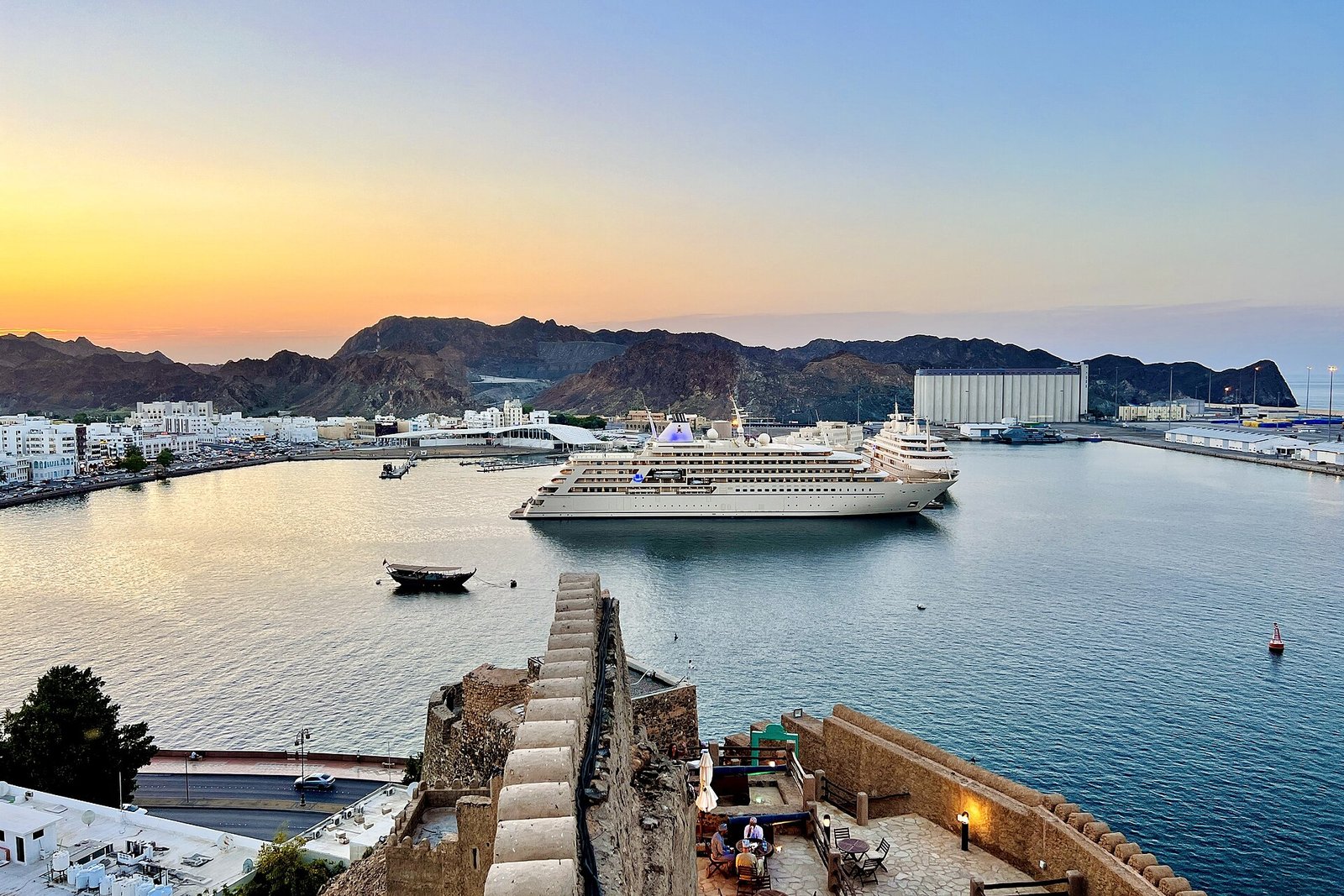 A cruise ship berthed in Muscat's Port Sultan Qaboos at sunset, with the Hajar Mountains behind.