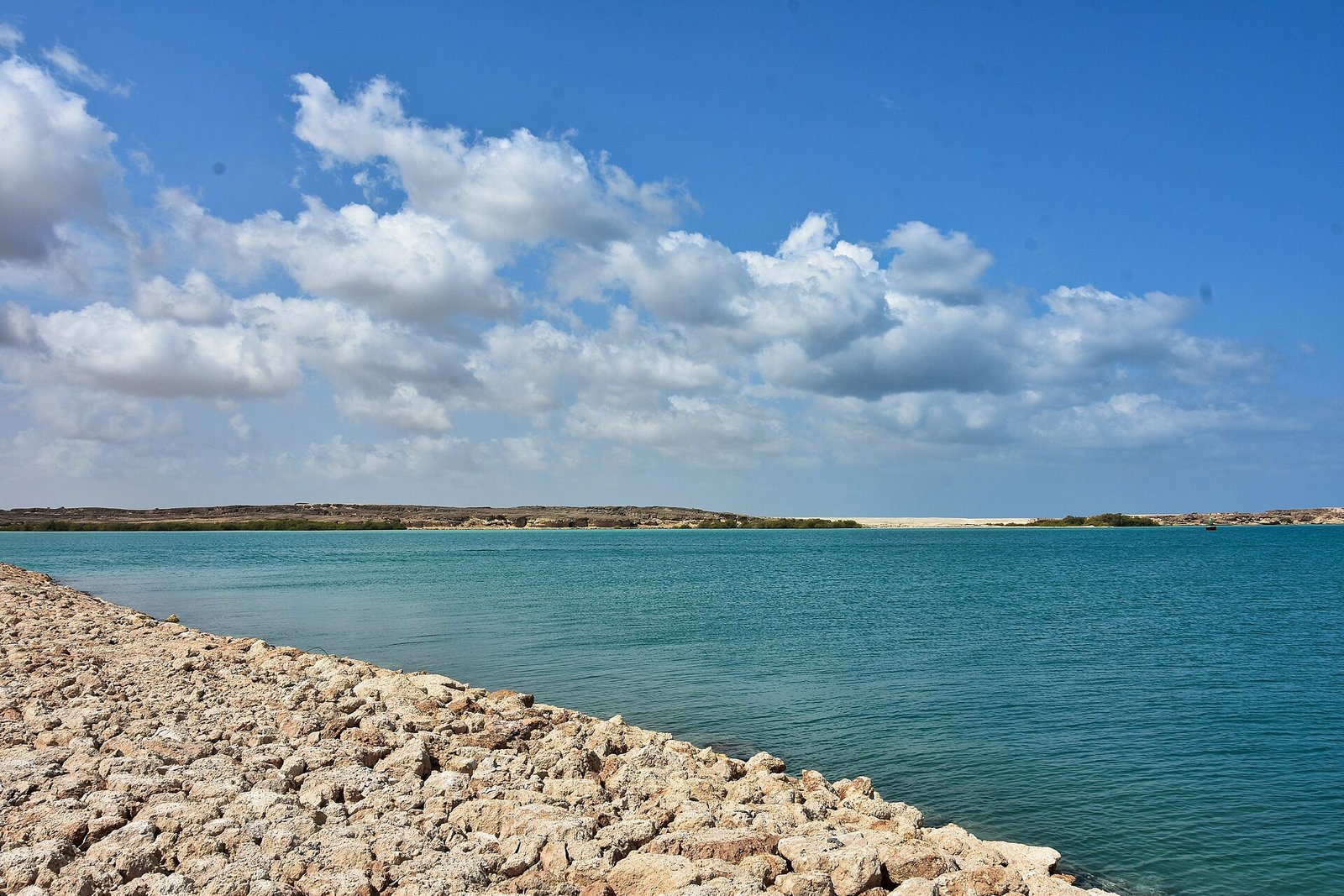 Turquoise Red Sea shallows and a coral-stone shore under white clouds at Farasan Island, Saudi Arabia.