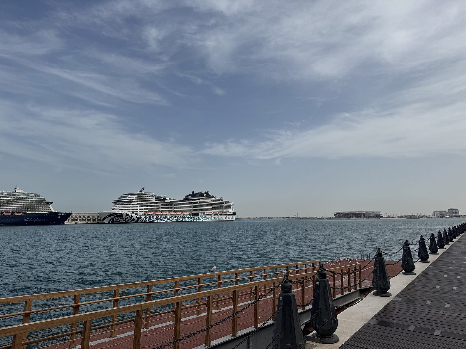 Cruise ships berthed at the port of Doha, seen across the calm water of Doha Bay from the waterfront promenade.