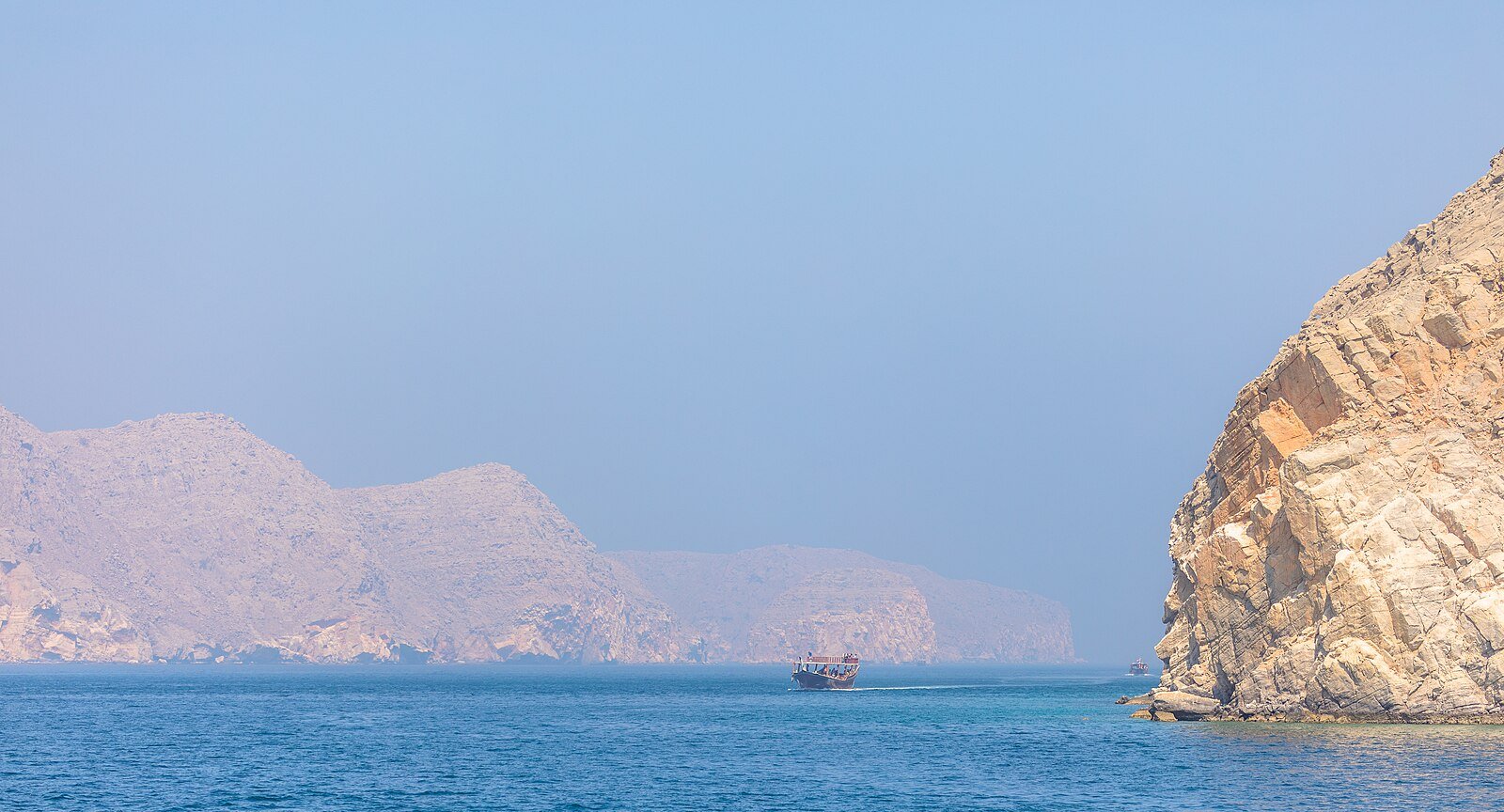 A dhow crossing the blue water of the Musandam fjords beneath sheer limestone cliffs, Oman.