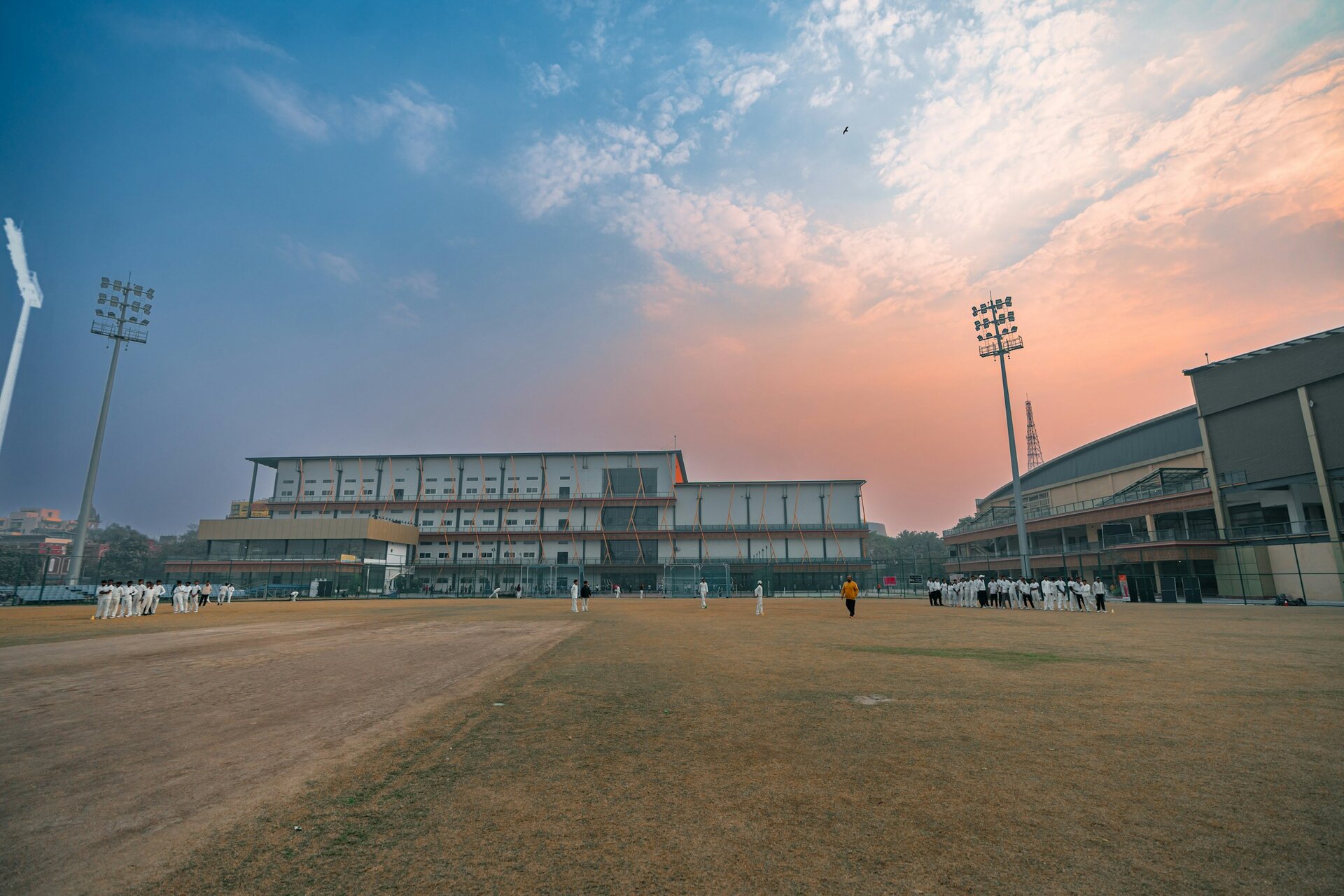 A cricket ground under floodlight towers at sunset, with squads in whites at practice