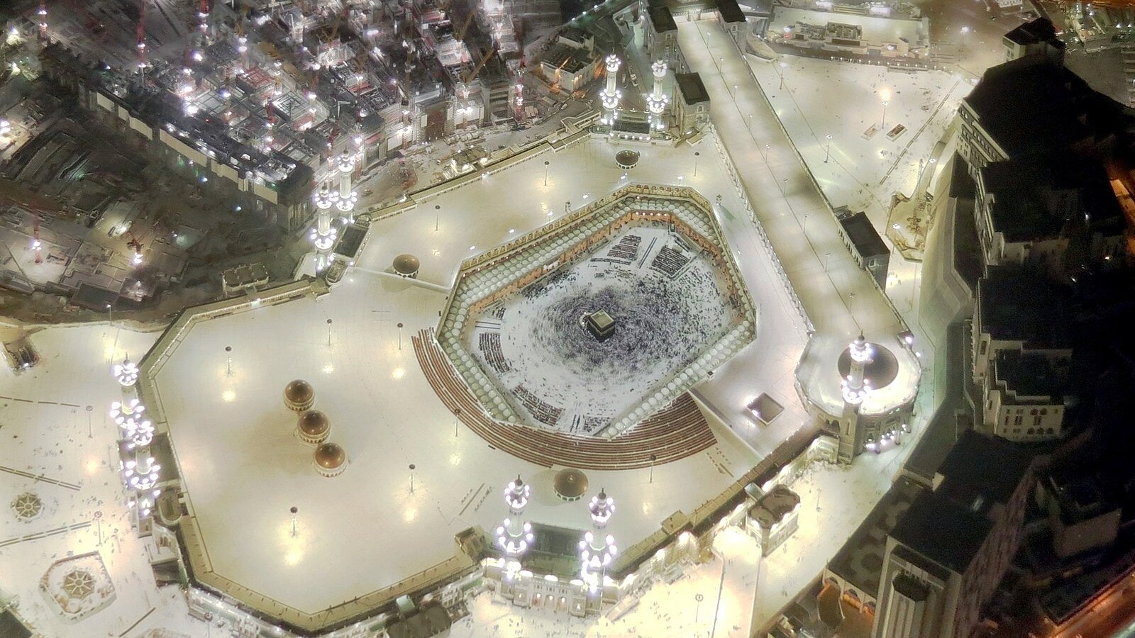 The illuminated courtyards and minarets of the Masjid al-Haram seen from high above at night, with the Kaaba at its heart.