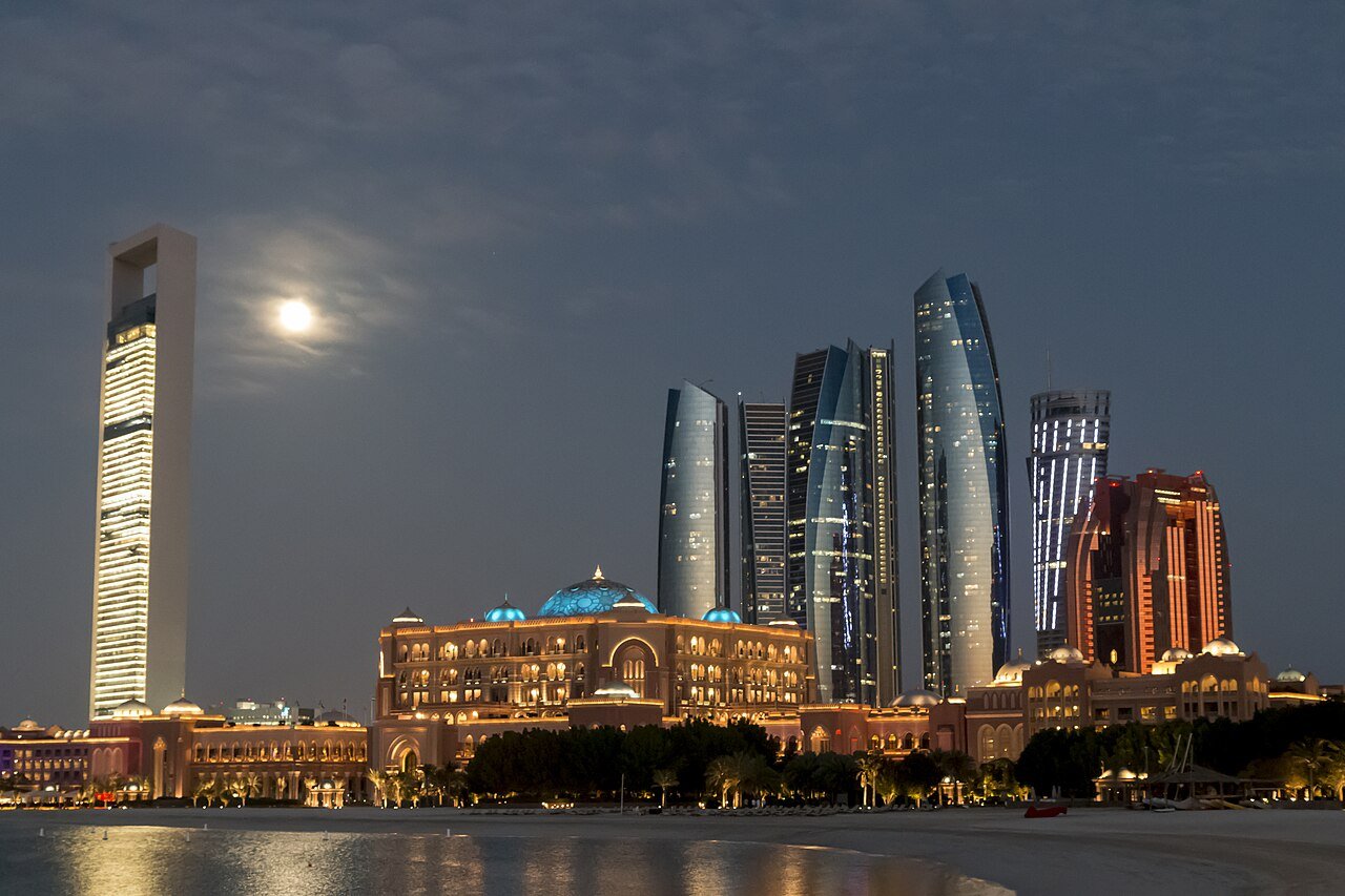 Emirates Palace and the Etihad Towers lit beneath a rising moon on the Abu Dhabi waterfront.