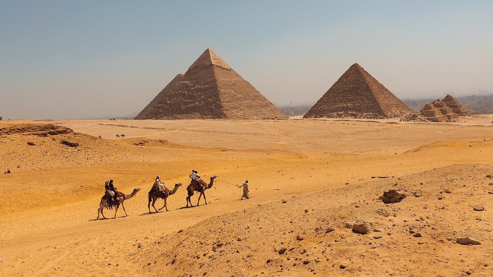 A camel caravan crossing the golden sands of the Giza Plateau before the pyramids of Khafre and Menkaure