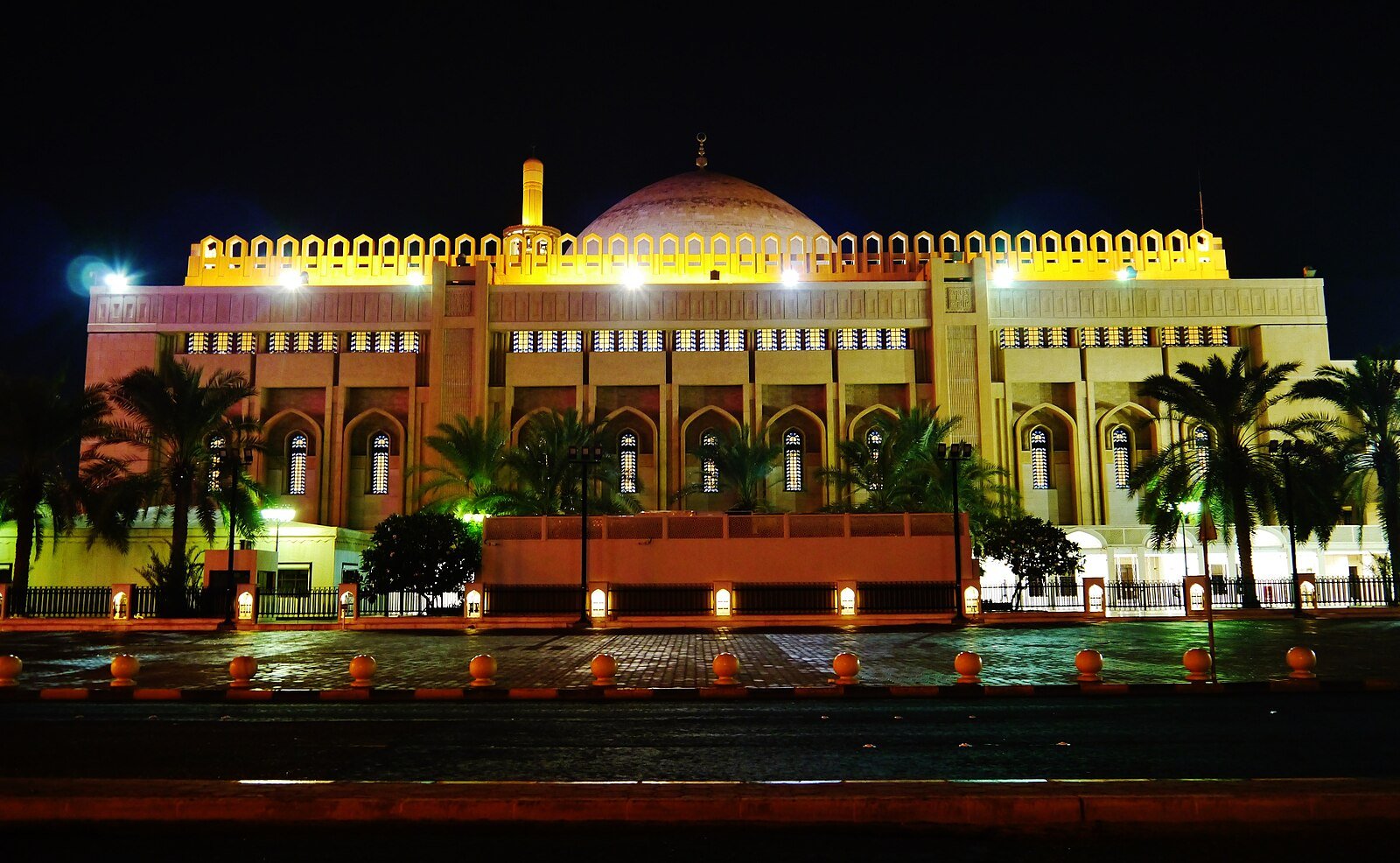 The Grand Mosque of Kuwait City glowing gold at night behind a row of date palms