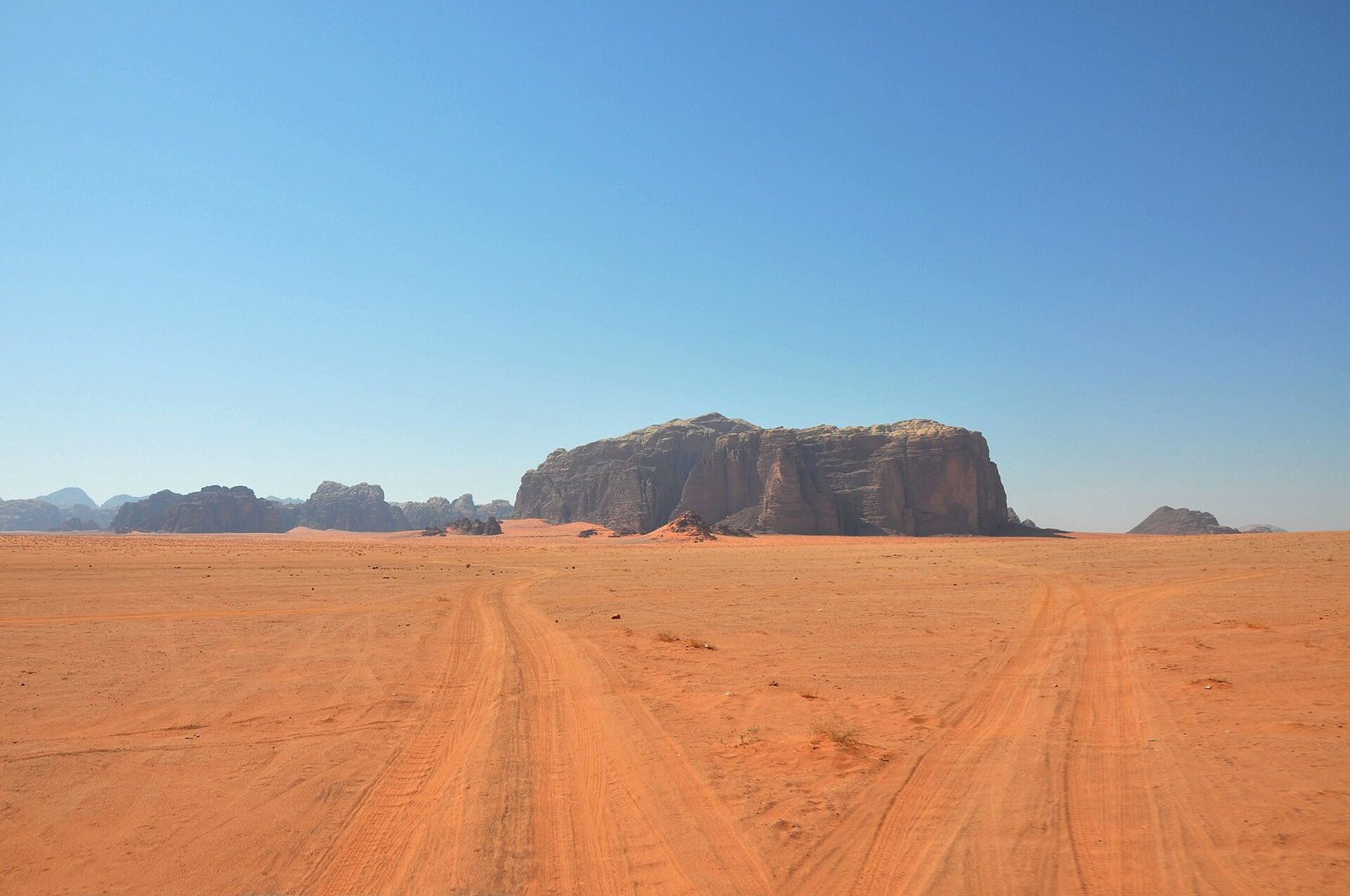 Jeep tracks cross the red sands of Wadi Rum toward a sandstone massif under a cloudless blue sky.