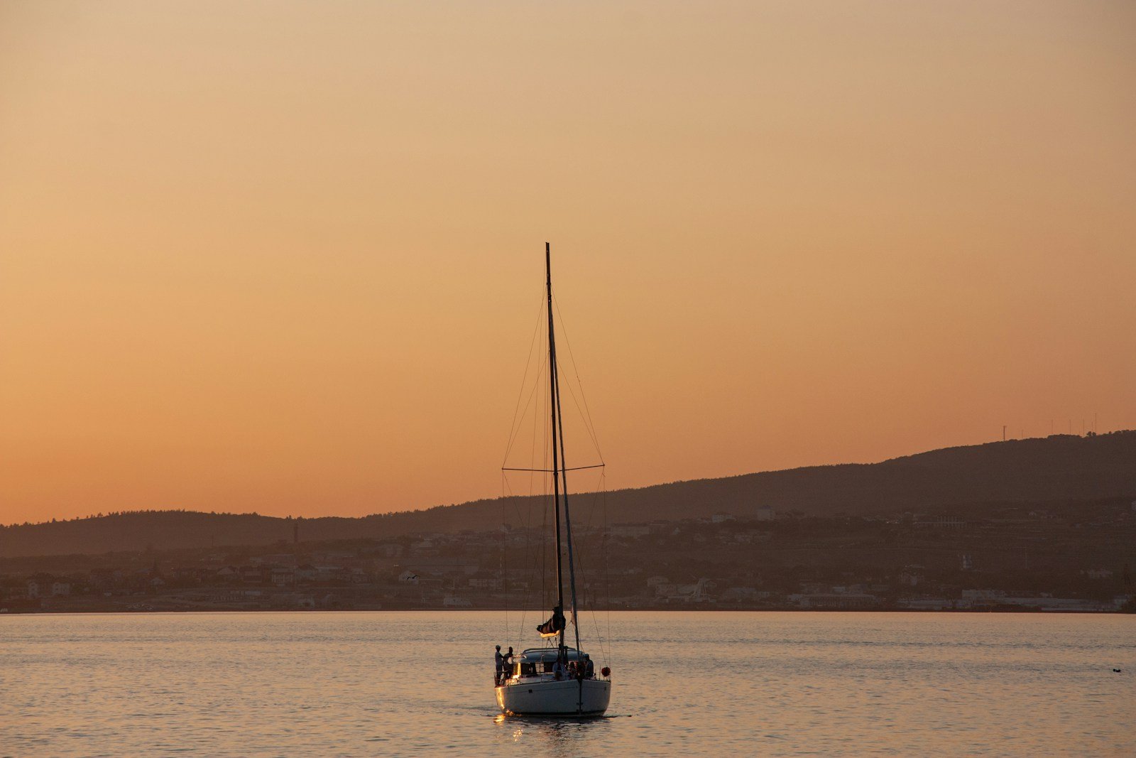 A sailing yacht rides at anchor on a glassy sea under an amber sunset sky.