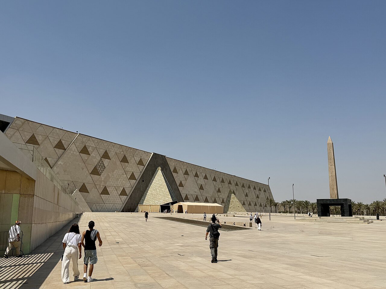 The triangular-patterned stone facade of the Grand Egyptian Museum and its obelisk under a clear Giza sky.