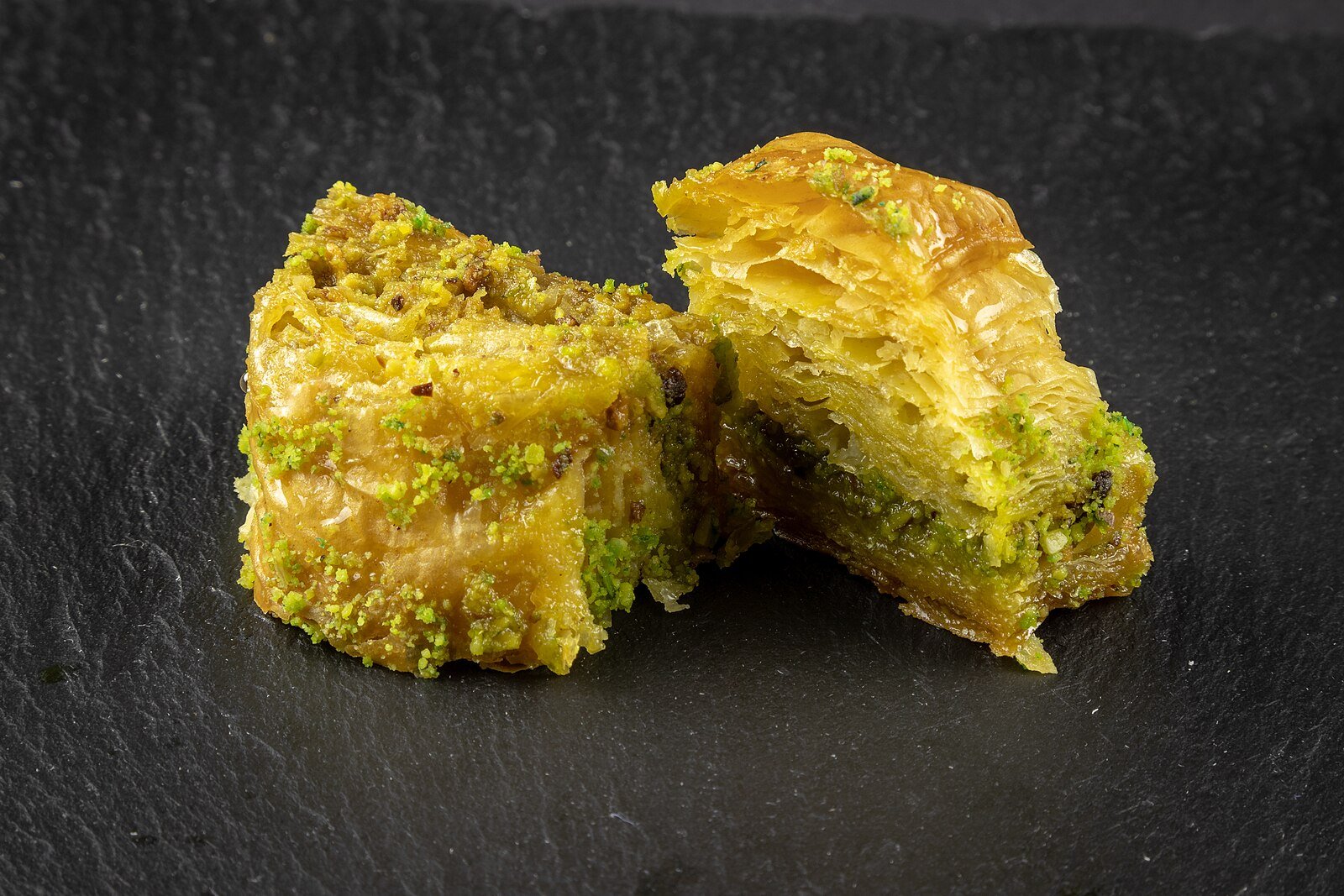 Layered pistachio baklava, glistening with syrup, on a dark slate.