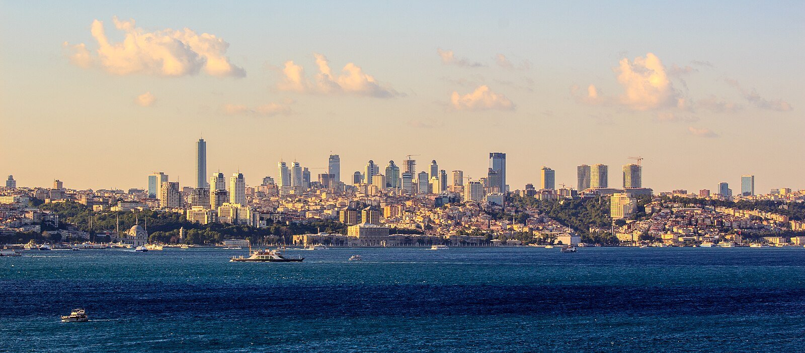 The modern skyline of Istanbul above the Bosphorus at sunset, with ferries crossing the water.