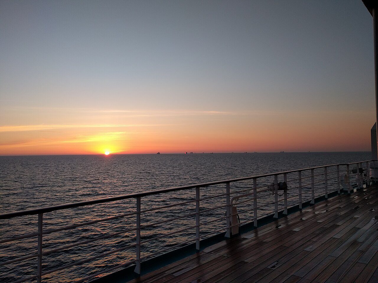 El sol poniéndose sobre un mar en calma, visto desde la cubierta de paseo de teca de un crucero.
