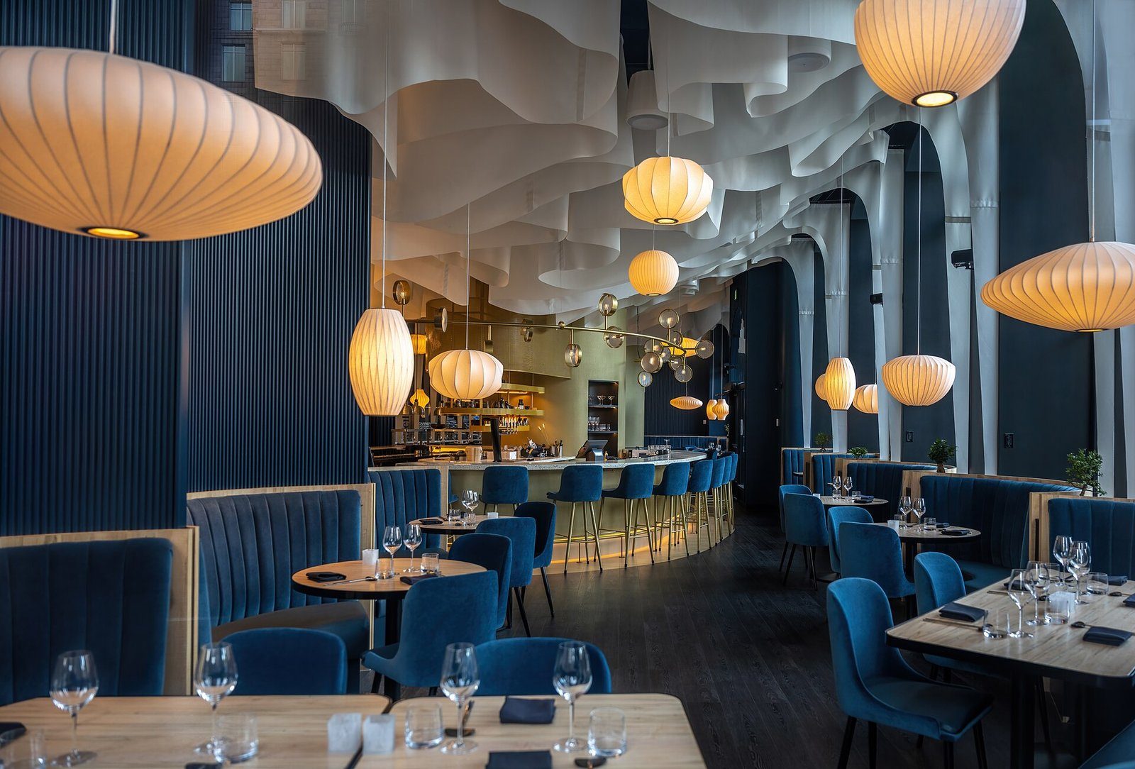 Un elegante comedor con banquetas de azul profundo, mesas puestas y farolillos de papel encendidos bajo un techo esculpido
