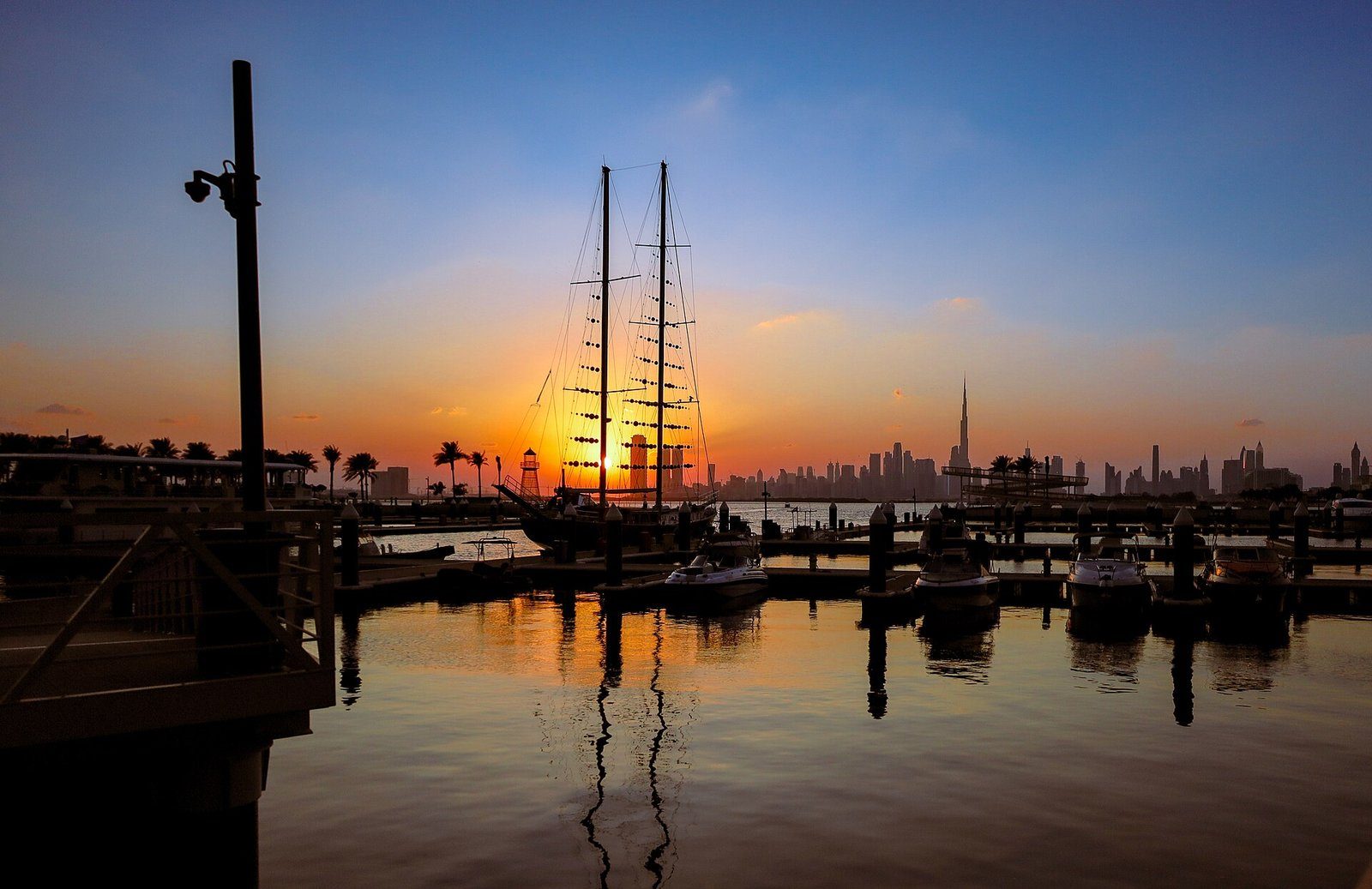 Los mástiles de un velero amarrado recortados contra un atardecer dorado sobre Dubai Creek Harbour, con el perfil urbano de Dubái al fondo.