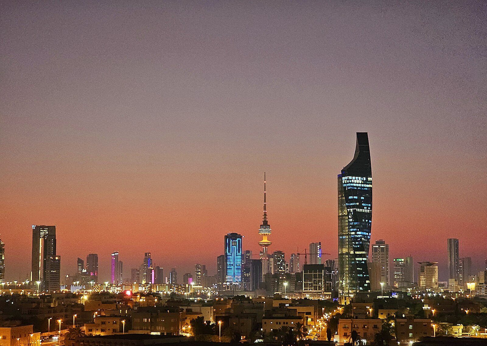 El perfil urbano de la ciudad de Kuwait al anochecer, con la Al Hamra Tower y la Liberation Tower iluminadas ante un cielo rosado.
