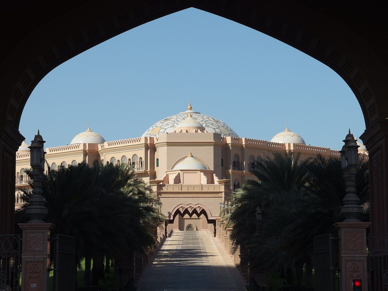Emirates Palace y sus cúpulas enmarcadas por un arco apuntado al final de una avenida flanqueada por palmeras, Abu Dabi.