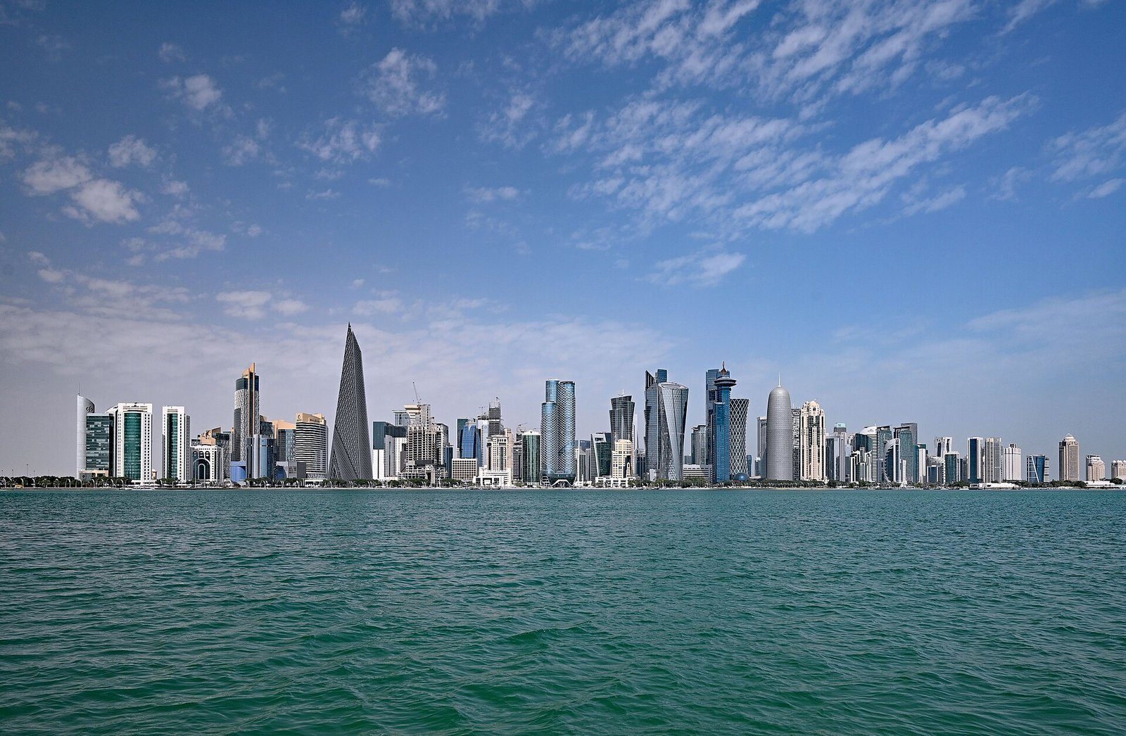 Las torres de West Bay en Doha alzándose sobre las aguas turquesas de la bahía de Doha bajo un cielo surcado de nubes.