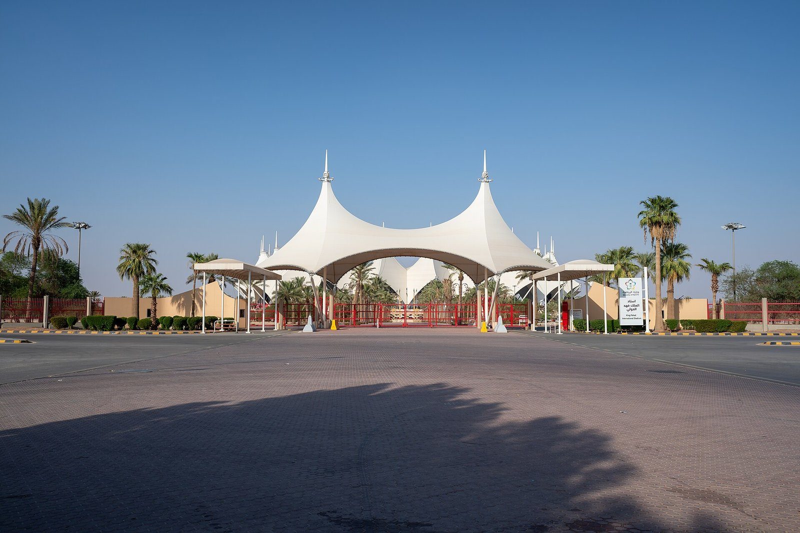 La blanca entrada con cubierta de lona del King Fahd International Stadium de Riad, flanqueada de palmeras bajo un cielo despejado.