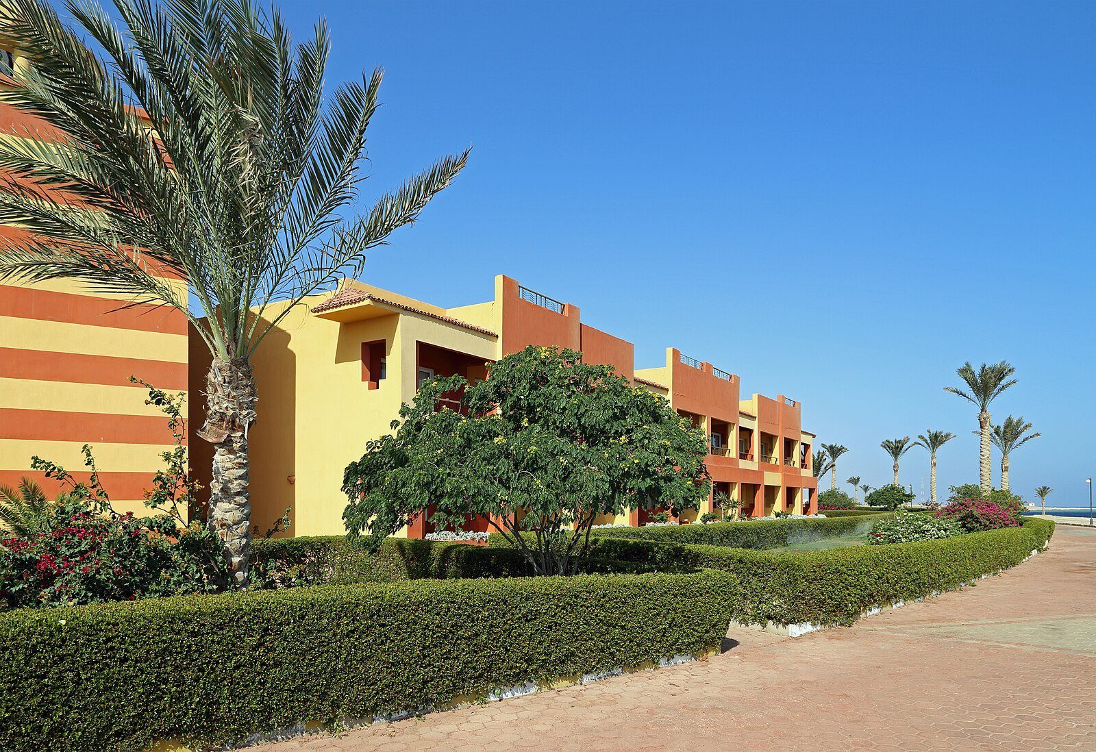 Jardines a la sombra de palmeras y los edificios con franjas ocres de un resort de playa en Abu Dabbab, Marsa Alam.