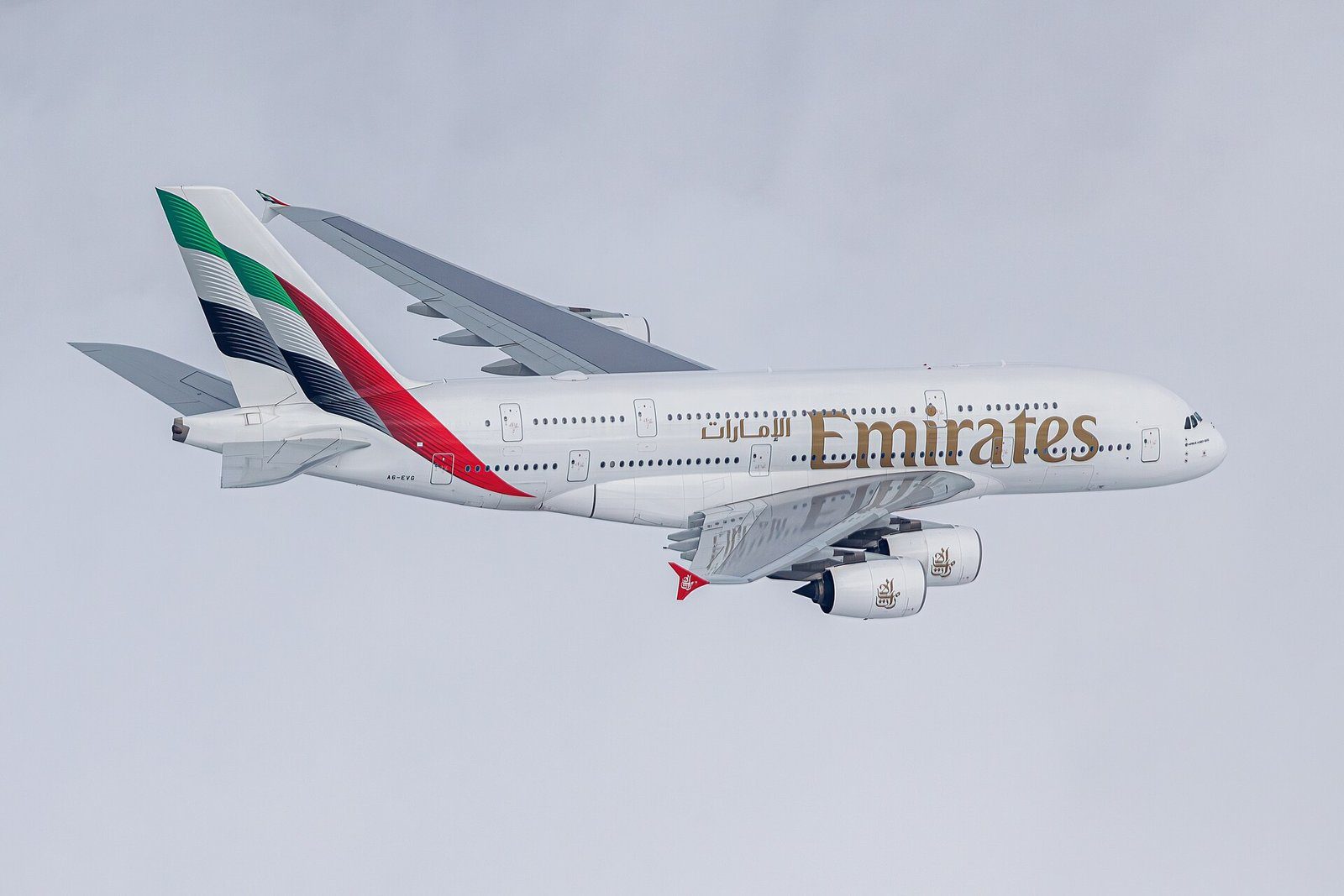 Un Airbus A380 de Emirates en vuelo, con su doble cubierta y la librea roja, verde y negra de la cola contra un cielo pálido.