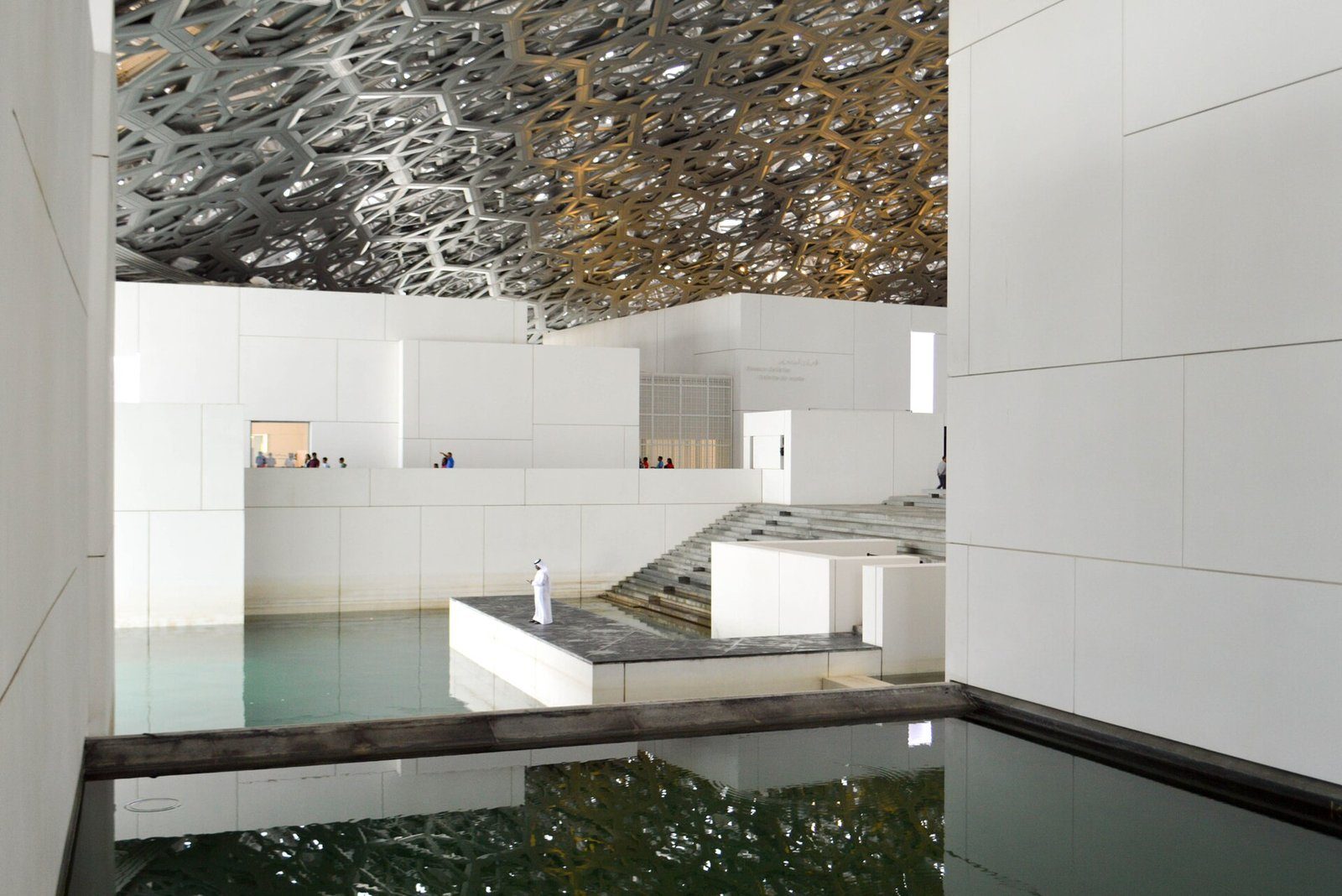 Galerías blancas y estanques reflectantes bajo la cúpula plateada y calada del Louvre Abu Dhabi.